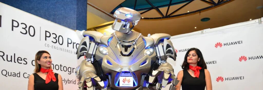 Titan the Robot stands with two models on either side, the Huawei logo on his chest and on a sign in the background.
