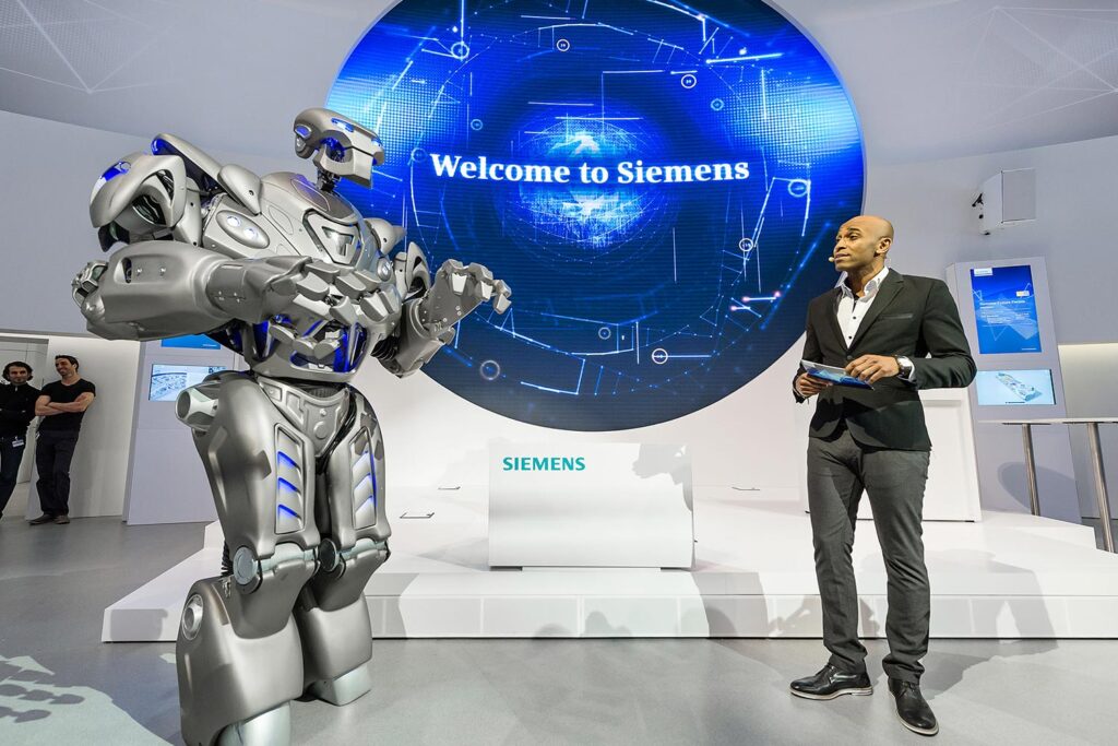 Titan the Robot and a presenter in front of a large futuristic digital display reading ‘Welcome to Siemens’.